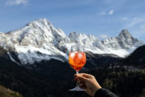Hand holding a Spritz cocktail against a backdrop of snow-covered Dolomites mountains, enjoying an Alpine aperitivo moment.