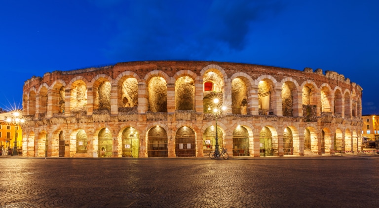 Verona Arena illuminated at night, showcasing its ancient Roman amphitheater structure against a blue evening sky.