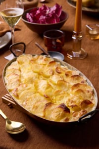 Potato dauphinois on a beautifully styled table with a tobacco-coloured tablecloth, amber candle, radicchio salad, and a glass of champagne, ready for an elegant autumn gathering.