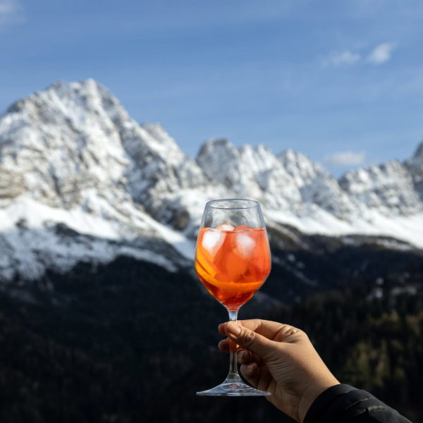 Hand holding a Spritz cocktail against a backdrop of snow-covered Dolomites mountains, enjoying an Alpine aperitivo moment.