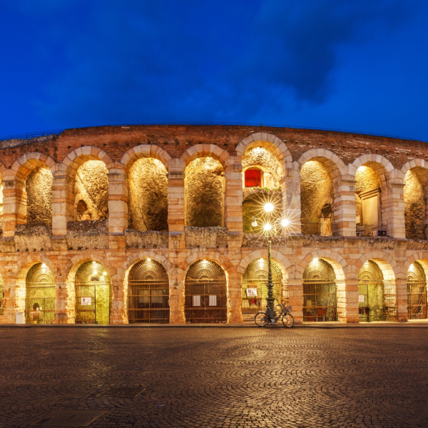 Verona Arena illuminated at night, showcasing its ancient Roman amphitheater structure against a blue evening sky.